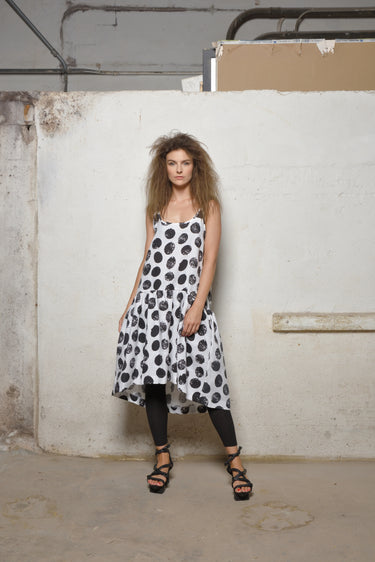 A female model wearing a spaghetti straps polka dots peplum dress with pockets, low neck line, asymmetrical hem, paired with Black leggings, black sandals against an industrial white background.