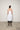 A female model wearing a spaghetti straps white peplum dress with strokes paint design on the side, pockets, low neck line, asymmetrical hem, paired with Black leggings, black sandals against an industrial white background.
