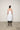 A female model wearing a spaghetti straps white peplum dress with strokes paint design on the side, pockets, low neck line, asymmetrical hem, paired with Black leggings, black sandals against an industrial white background.