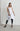 A female model wearing a spaghetti straps white peplum dress with strokes paint design on the side, pockets, low neck line, asymmetrical hem, paired with Black leggings, black sandals against an industrial white background.