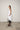 A female model wearing a spaghetti straps white peplum dress with strokes paint design on the side, pockets, low neck line, asymmetrical hem, paired with Black leggings, black sandals against an industrial white background.