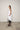 A female model wearing a spaghetti straps white peplum dress with strokes paint design on the side, pockets, low neck line, asymmetrical hem, paired with Black leggings, black sandals against an industrial white background.