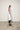 A female model wearing a spaghetti straps white peplum dress with strokes paint design on the side, pockets, low neck line, asymmetrical hem, paired with Black leggings, black sandals against an industrial white background.