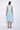 A woman wearing a light blue sleeveless dress with a square neckline, an asymmetrical, layered design with diagonal seam lines, prominent gathered, pockets, irregular hemline, pairing with neutral tones sandals over a white background.