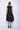 A woman wearing a black sleeveless dress with a square neckline, an asymmetrical, layered design with diagonal seam lines, prominent gathered, pockets, irregular hemline, pairing with black sandals over a white background.