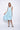A woman wearing a light blue sleeveless dress with a square neckline, an asymmetrical, layered design with diagonal seam lines, prominent gathered, pockets, irregular hemline, pairing with neutral tones sandals over a white background.