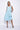 A woman wearing a light blue sleeveless dress with a square neckline, an asymmetrical, layered design with diagonal seam lines, prominent gathered, pockets, irregular hemline, pairing with neutral tones sandals over a white background.