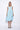 A woman wearing a light blue sleeveless dress with a square neckline, an asymmetrical, layered design with diagonal seam lines, prominent gathered, pockets, irregular hemline, pairing with neutral tones sandals over a white background.