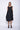 A woman wearing a black sleeveless dress with a square neckline, an asymmetrical, layered design with diagonal seam lines, prominent gathered, pockets, irregular hemline, pairing with black sandals over a white background.