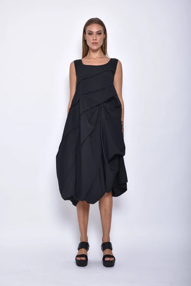 A woman wearing a black sleeveless dress with a square neckline, an asymmetrical, layered design with diagonal seam lines, prominent gathered, pockets, irregular hemline, pairing with black sandals over a white background.