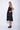 A woman wearing a black sleeveless dress with a square neckline, an asymmetrical, layered design with diagonal seam lines, prominent gathered, pockets, irregular hemline, pairing with black sandals over a white background.