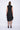 A woman wearing a sleeveless black dress with a sporty, utilitarian design. The dress includes a hood, a front zipper closure, and drawstring details at the waist and hem, allowing for adjustable ruching. The silhouette is voluminous and sculptural, particularly around the hemline, which forms a balloon-like shape, chunky black platform sandals. The background is plain white.