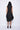 A woman wearing a sleeveless black dress with a sporty, utilitarian design. The dress includes a hood, a front zipper closure, and drawstring details at the waist and hem, allowing for adjustable ruching. The silhouette is voluminous and sculptural, particularly around the hemline, which forms a balloon-like shape, chunky black platform sandals. The background is plain white.