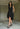A woman wearing a sleeveless black dress with a sporty, utilitarian design. The dress includes a hood, a front zipper closure, and drawstring details at the waist and hem, allowing for adjustable ruching. The silhouette is voluminous and sculptural, particularly around the hemline, which forms a balloon-like shape, chunky black platform sandals. The background is plain white.