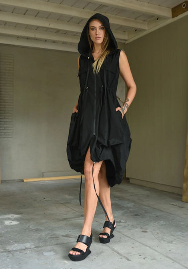 A woman wearing a sleeveless black dress with a sporty, utilitarian design. The dress includes a hood, a front zipper closure, and drawstring details at the waist and hem, allowing for adjustable ruching. The silhouette is voluminous and sculptural, particularly around the hemline, which forms a balloon-like shape, chunky black platform sandals. The background is plain white.