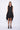A woman wearing a sleeveless black dress with a sporty, utilitarian design. The dress includes a hood, a front zipper closure, and drawstring details at the waist and hem, allowing for adjustable ruching. The silhouette is voluminous and sculptural, particularly around the hemline, which forms a balloon-like shape, chunky black platform sandals. The background is plain white.