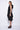 A woman wearing a sleeveless black dress with a sporty, utilitarian design. The dress includes a hood, a front zipper closure, and drawstring details at the waist and hem, allowing for adjustable ruching. The silhouette is voluminous and sculptural, particularly around the hemline, which forms a balloon-like shape, chunky black platform sandals. The background is plain white.