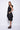 A woman wearing a sleeveless black dress with a sporty, utilitarian design. The dress includes a hood, a front zipper closure, and drawstring details at the waist and hem, allowing for adjustable ruching. The silhouette is voluminous and sculptural, particularly around the hemline, which forms a balloon-like shape, chunky black platform sandals. The background is plain white.