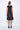 A model wearing a sleeveless, black dress with a black abstract design at the bottom. The dress is knee-length and the model is wearing black sandals.