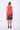A model wearing a sleeveless, red dress with a black abstract design at the bottom. The dress is knee-length and the model is wearing black sandals.