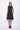 A model wearing a sleeveless, black dress with a black abstract design at the bottom. The dress is knee-length and the model is wearing black sandals.