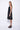 A model wearing a sleeveless, black dress with a black abstract design at the bottom. The dress is knee-length and the model is wearing black sandals.