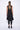 A woman wearing a sleeveless black sheer dress with a scoop neckline. The dress has a voluminous, sculptural silhouette with gathered and ruched detailing around the hemline, paired with black leggings and chunky black open-toe heels sandals over a white background.