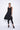 A  woman wearing a sleeveless black sheer dress with a scoop neckline. The dress has a voluminous, sculptural silhouette with gathered and ruched detailing around the hemline, paired with black leggings and chunky black open-toe heels sandals over a white background.