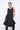 A woman wearing a sleeveless black sheer dress with a scoop neckline. The dress has a voluminous, sculptural silhouette with gathered and ruched detailing around the hemline, paired with black leggings and chunky black open-toe heels sandals over a white background.