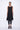 A woman wearing a sleeveless black sheer dress with a scoop neckline. The dress has a voluminous, sculptural silhouette with gathered and ruched detailing around the hemline, paired with black leggings and chunky black open-toe heels sandals over a white background.
