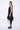A woman wearing a sleeveless black sheer dress with a scoop neckline. The dress has a voluminous, sculptural silhouette with gathered and ruched detailing around the hemline, paired with black leggings and chunky black open-toe heels sandals over a white background.