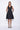 A model wearing a black sleeveless knee-length dress with a v-neckline, center zipper and pockets, standing against a white background.