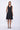 A model wearing a black sleeveless knee-length dress with a v-neckline, center zipper and pockets, standing against a white background.