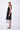 A model wearing a black sleeveless knee-length dress with a v-neckline, center zipper and pockets, standing against a white background.