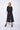 A female model wearing black pants and a matching black top, standing against a white background.