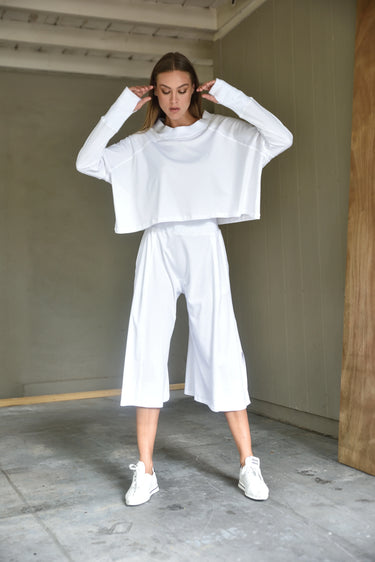 A female model wearing a white turtleneck top and white long pants, standing in an indoor setting.