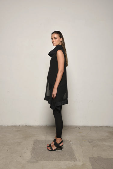 A woman modeling a sleeveless black tunic with a draped cowl neck and Asymmetrical hem, paired with black leggings.