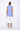 A female model wearing a blue sleeveless asymmetric top with a longer back length and paired with white pants.