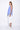 A female model wearing a blue sleeveless asymmetric top with a longer back length and paired with white pants.