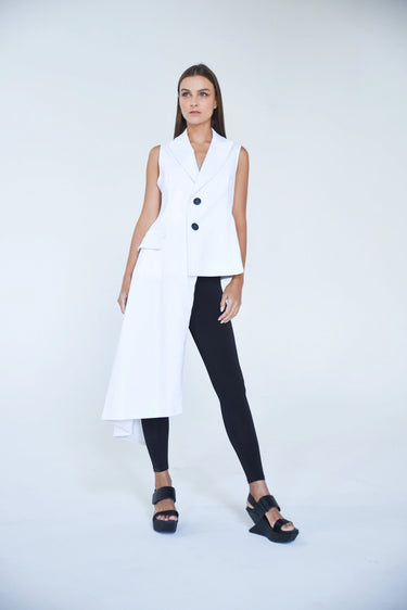 A model wearing an asymmetrical white vest with no sleeves, paired with black pants and black sandals.