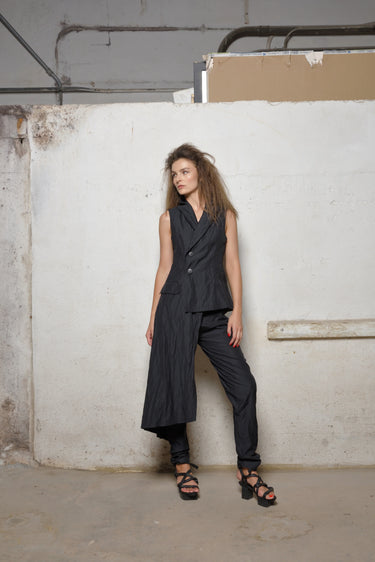 A woman standing against a white backdrop wearing an asymmetrical sleeveless black vest and black pants.