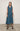 A woman standing against a white backdrop wearing an asymmetrical sleeveless Teal vest and Teal cropped pants.