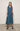 A woman standing against a white backdrop wearing an asymmetrical sleeveless Teal vest and Teal cropped pants.