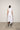 A woman standing against a white backdrop wearing an asymmetrical sleeveless White vest and White cropped with a side split pants.