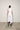 A woman standing against a white backdrop wearing an asymmetrical sleeveless White vest and White cropped with a side split pants.