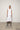 A woman standing against a white backdrop wearing an asymmetrical sleeveless White vest and White cropped with a side split pants.