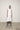 A woman standing against a white backdrop wearing an asymmetrical sleeveless White vest and White cropped with a side split pants.