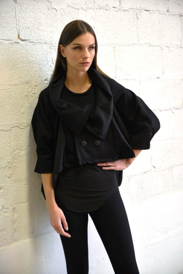 A woman wearing a black cropped jacket with 3/4 sleeves outfit standing against a white brick wall.