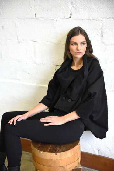 Woman wearing a black coat sitting on a wooden barrel against a white brick wall.