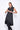 A woman wearing a sleeveless black trench-style Vest with a double-breasted button front, notched lapel collar and flap pockets on either side of the waist, asymmetrical hem line, paired with black leggings and chunky black open-toe heels over a white background.